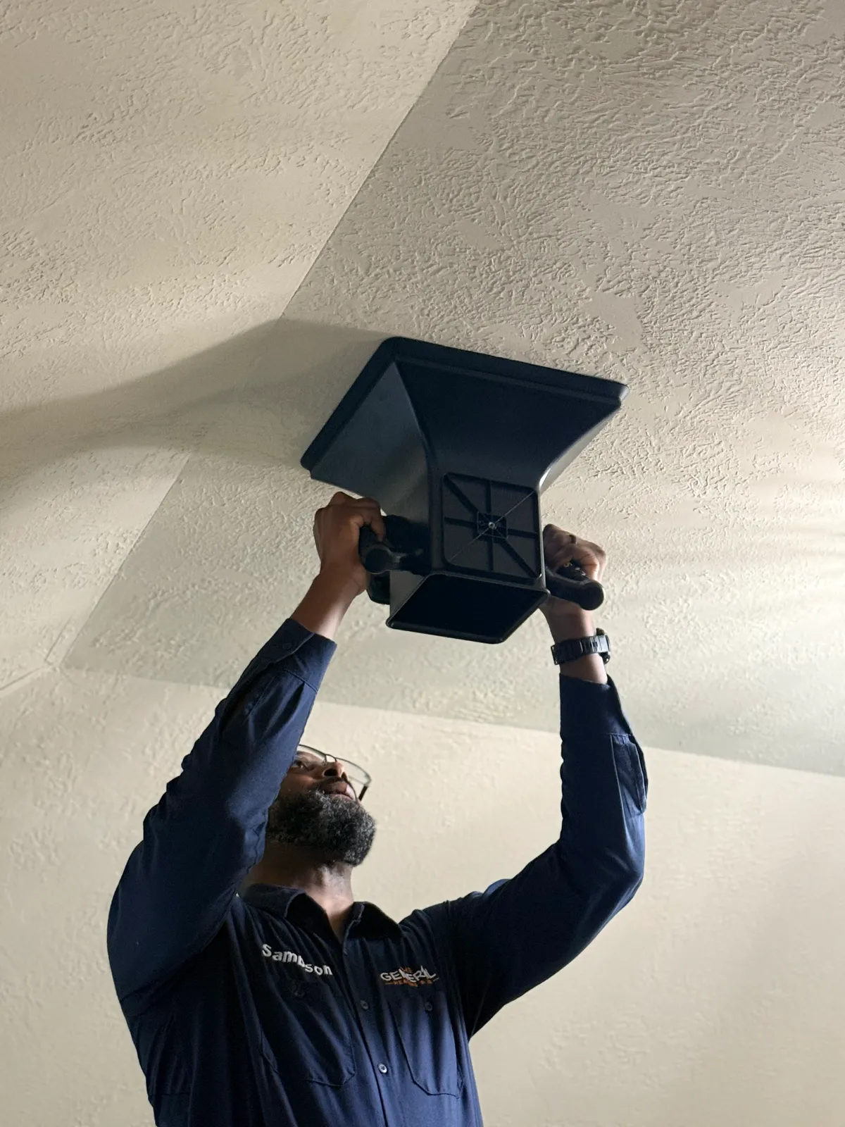 A technician from The General holds an airflow capture hood up to a ceiling supply register to measure airflow, in a navy branded uniform shirt.