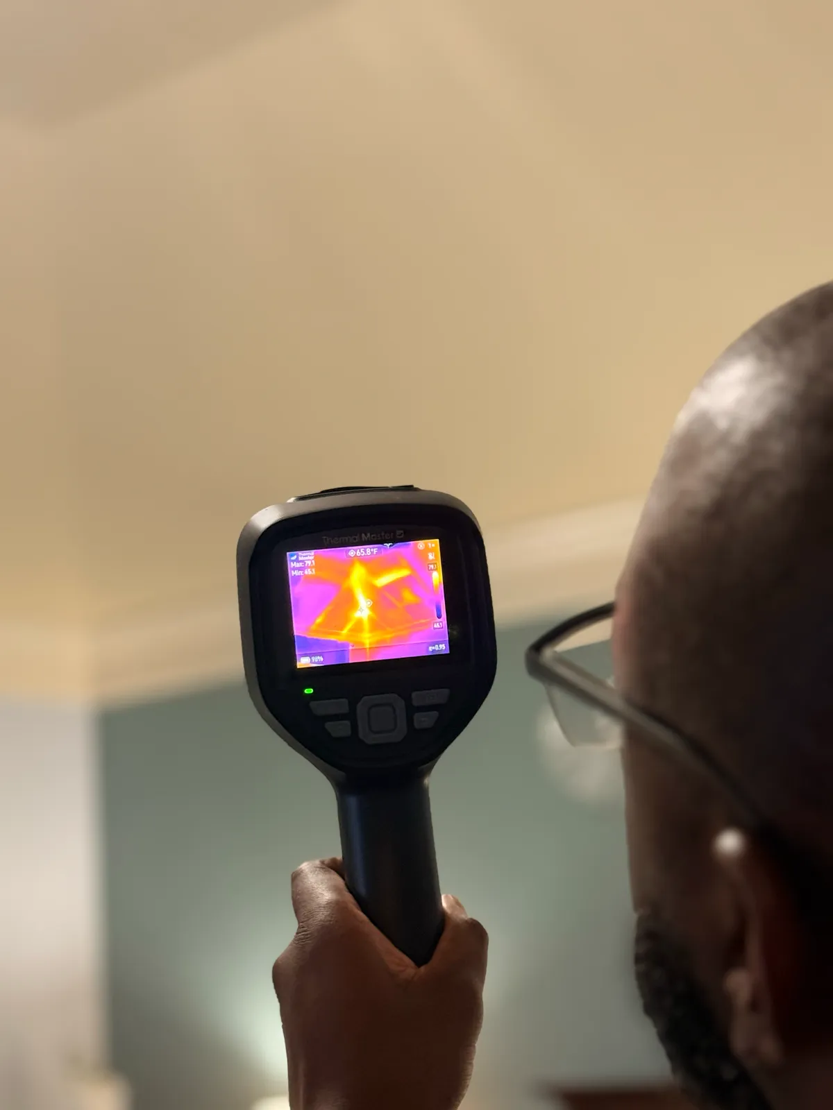 A technician aims a handheld thermal imaging camera at a ceiling register, the heat pattern around the vent visible on the camera screen.